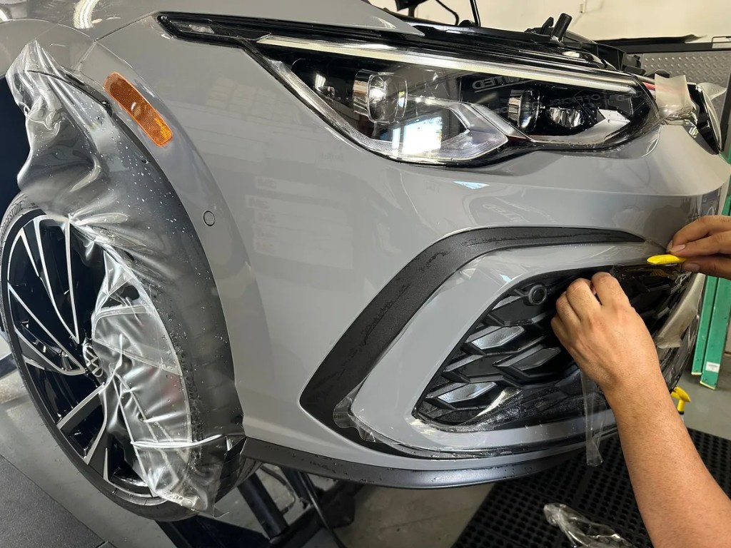 OCD installer wrapping SunTek paint protection film around the front bumper of a gray sports sedan